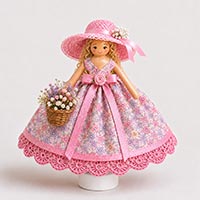 Finished handmade doll pin doll wearing a floral fabric dress trimmed with pink crocheted lace, a wide-brim pink abaca sun hat decorated with baby's breath flowers and satin ribbon, curly blonde hair, and holding a small basket of pastel flowers and baby's breath.