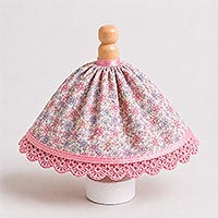 Instructional image showing the gathered fabric skirt positioned on a painted doll pin, with the top edge of the skirt covered with narrow pink satin ribbon and placed approximately 1/2 inch below the doll's neck to form the dress skirt.