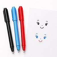 Three fine-tip markers in black, light blue, and red beside a sheet of paper with two practice doll face features, showing eyes, eyebrows, noses, and smiles for doll face painting.